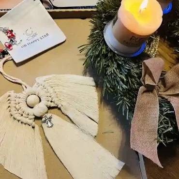 Handgefertigter Makrame-Engel aus naturfarbener Baumwolle, neben einer brennenden Kerze und einem Adventskranz arrangiert.