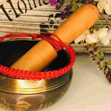 Handgefertigtes rotes tibetisches Armband mit traditioneller Stickerei, auf einer Klangschale mit Holzklöppel und Blüten im Hintergrund präsentiert.