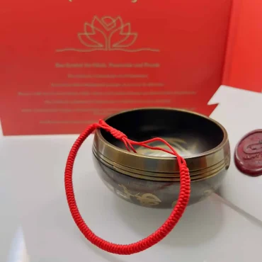 Rotes tibetisches Glücksarmband auf einer Klangschale, mit rotem Hintergrund und goldener Schrift – Symbol für Glück, Positivität und Freude.